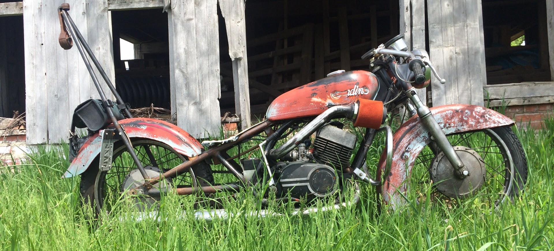 A patinated rigid-frame bobber standing in tall grass against a weathered barn