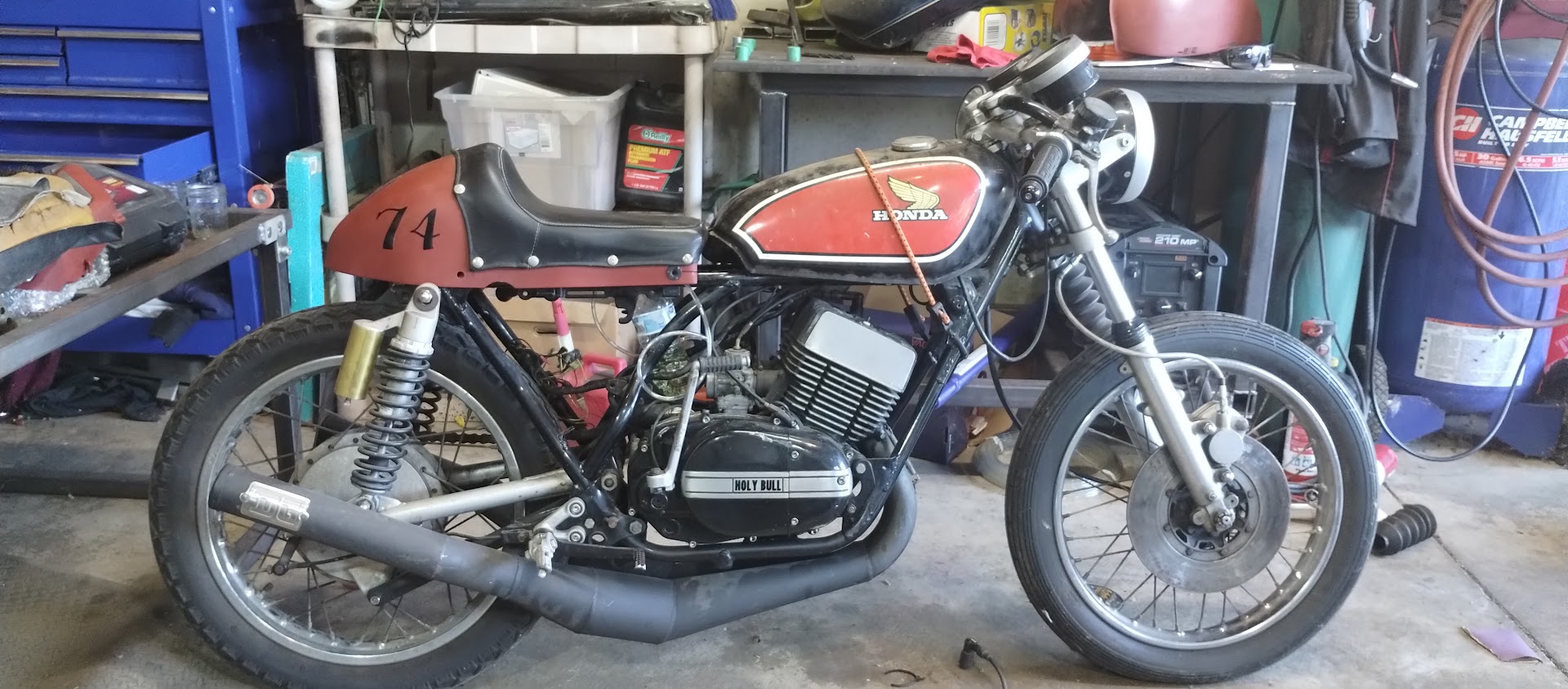 A Honda cafe racer with a number 74 tail section and Holy Bull engine cover, in the shop