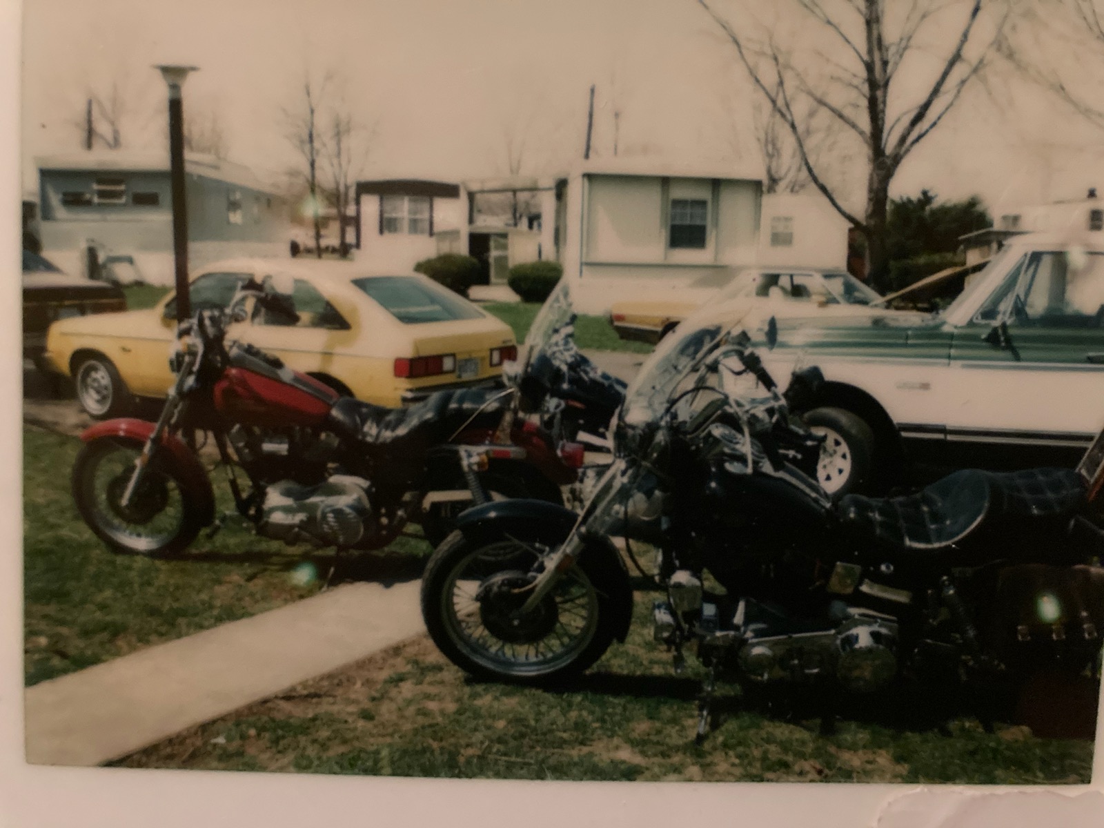 A faded snapshot of three motorcycles and two cars parked outside trailers