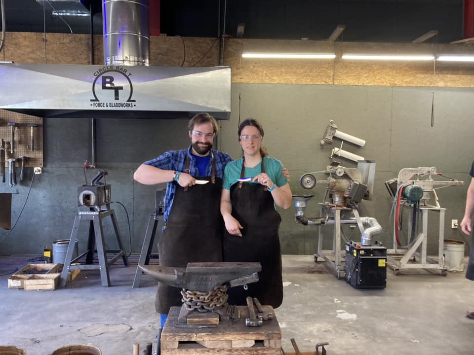 Two smiths in leather aprons holding finished knife blades in front of an anvil