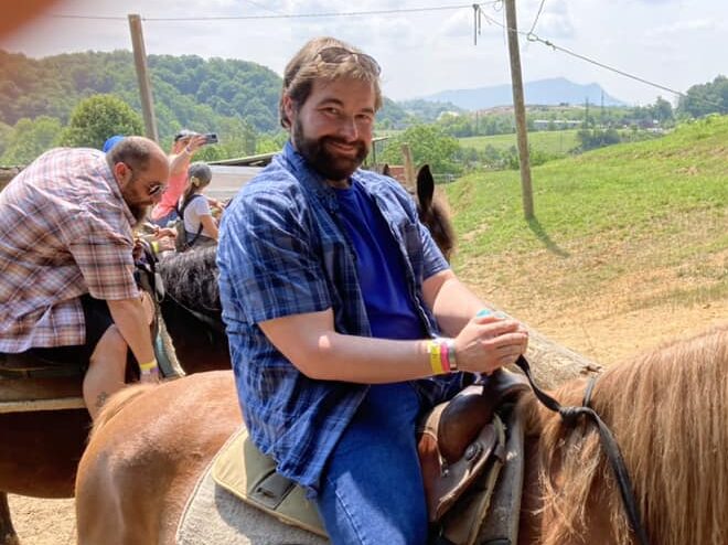 Greg, in a blue plaid shirt, sitting on horseback on a green hillside